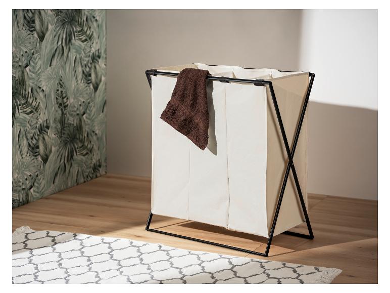 Een opvouwbare wasmand met drie compartimenten, van beige stof en een zwart frame, staat op een houten vloer.