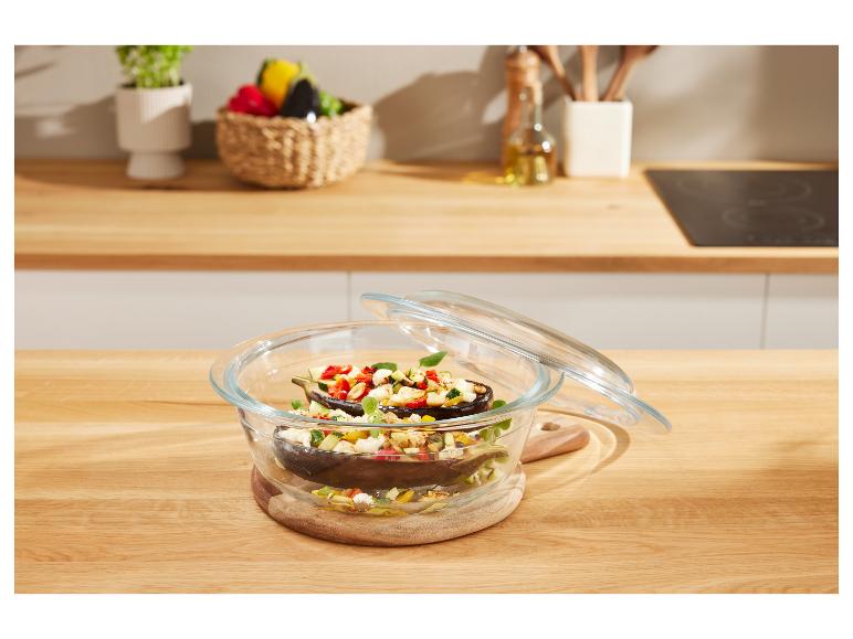 Glazen schaal met gevulde aubergines en glazen deksel op een houten tafel.