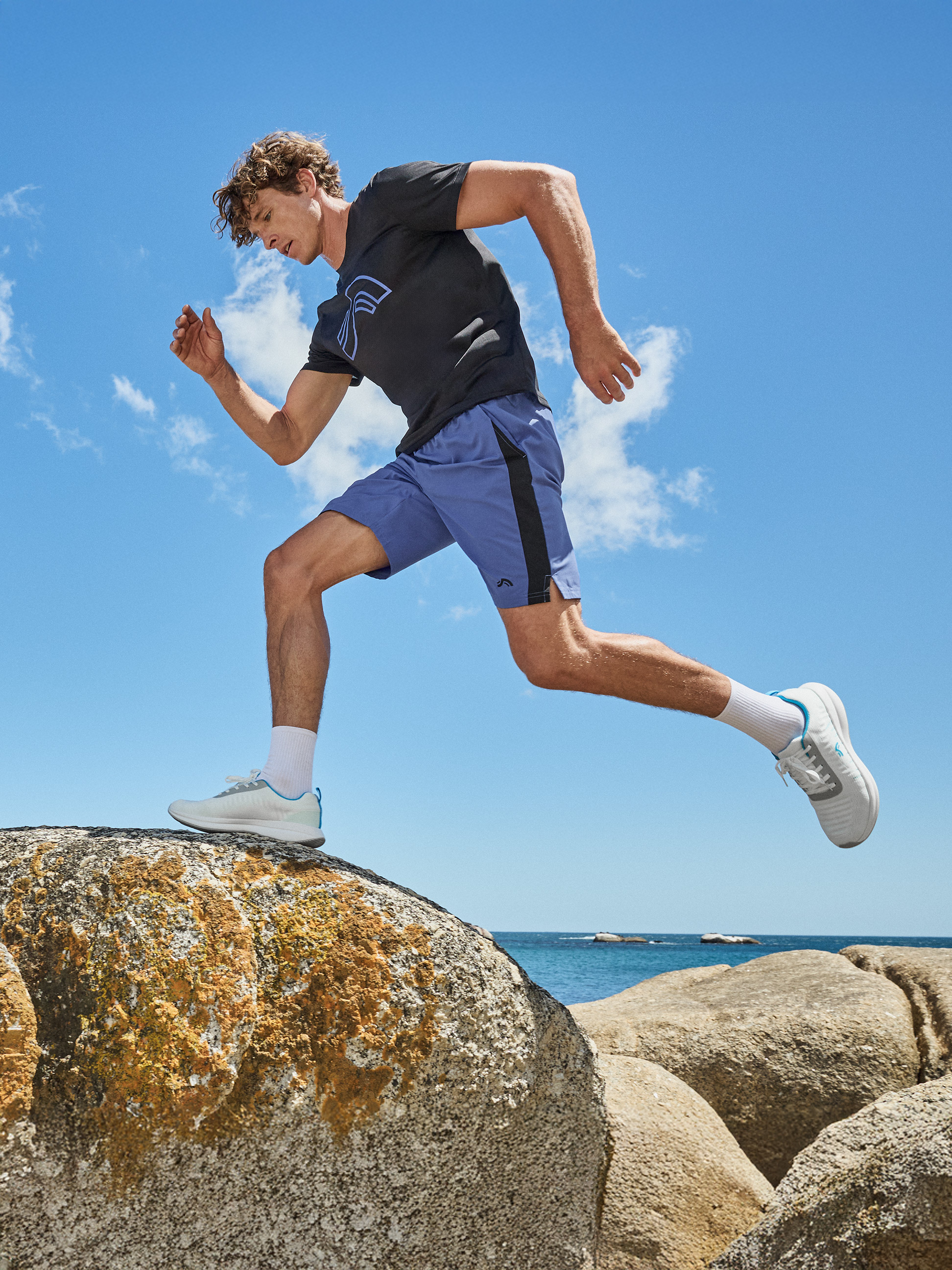 Man in zwart T-shirt en blauwe korte broek rent over rotsen aan het strand.