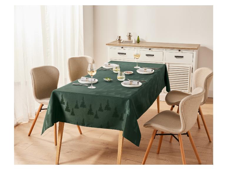 Een donkergroen tafelkleed met kerstboomprint, gedekt met bestek, glazen en servetten.