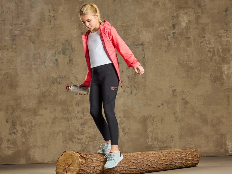 Een meisje draagt zwarte leggings met een roze jas en witte sneakers op een boomstam.