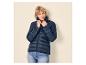 Vrouw in een donkerblauwe gewatteerde jas en jeans, met kort blond haar, kijkt naar rechts.