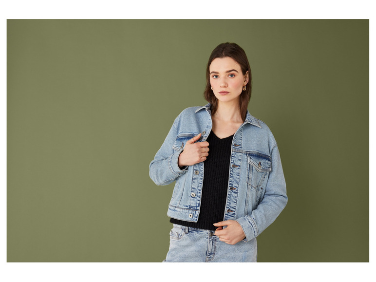Een vrouw in een denim jack en spijkerbroek poseert voor een groene achtergrond.