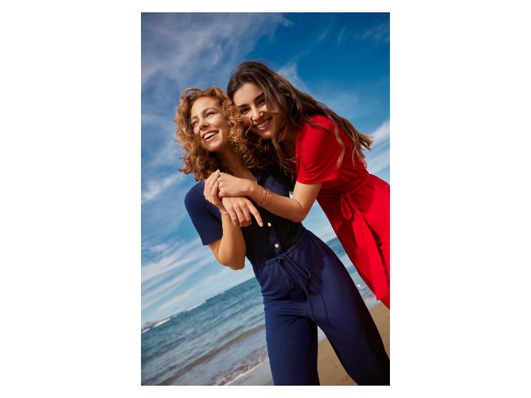 Twee lachende vrouwen in jumpsuits op het strand, één in marineblauw, één in rood.