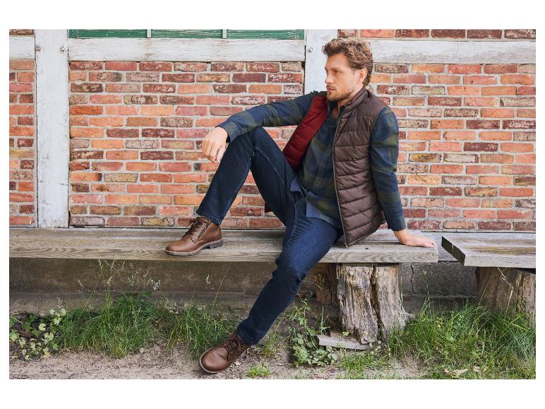 Man in bruin vest, geruit overhemd en jeans zittend op een houten bank.