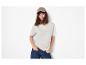 Vrouw in beige t-shirt, blauwe jeans en bruine pet, handen in zakken.