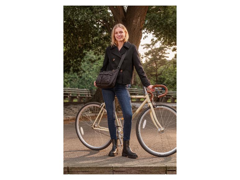Vrouw in zwarte peacoat en jeans met een zwarte schoudertas, staand naast een fiets.