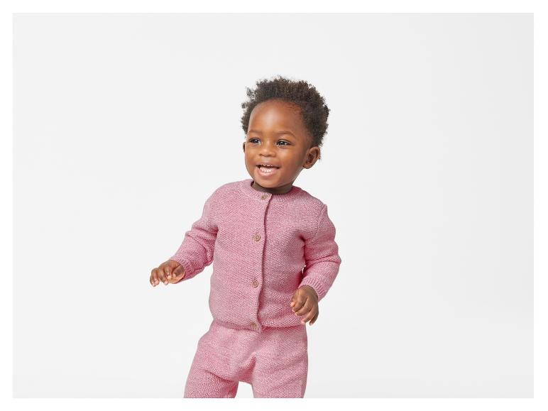 Een peuter lacht in een roze gebreid vest en broek.