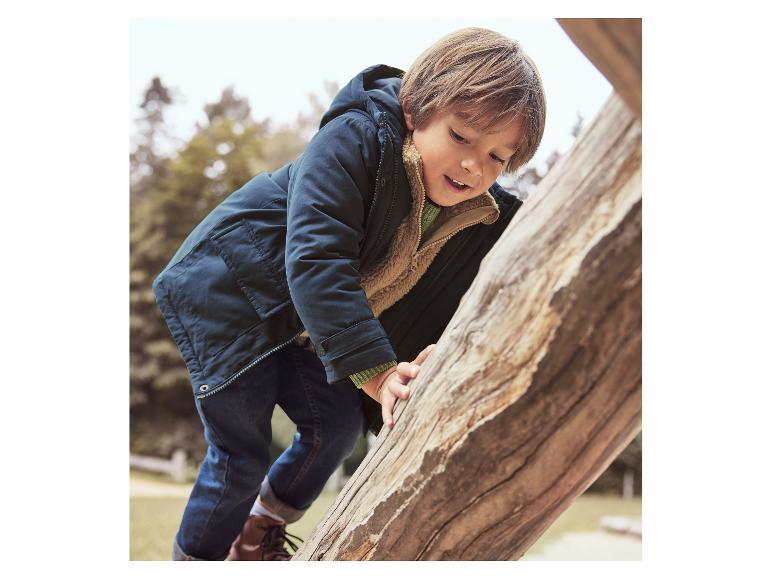 Een jongen in een donkerblauwe jas en jeans klimt op een boomstam.