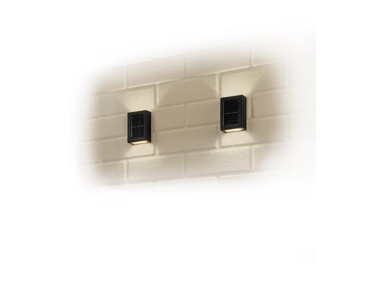Twee zwarte, rechthoekige buitenlampen met zonnepanelen, gemonteerd op een witte bakstenen muur.