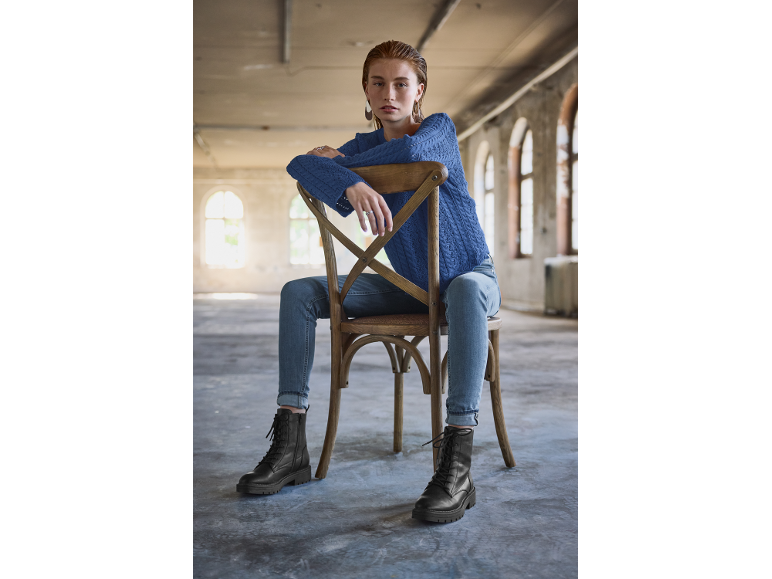 Vrouw in blauwe trui en zwarte laarzen zit op een houten stoel.