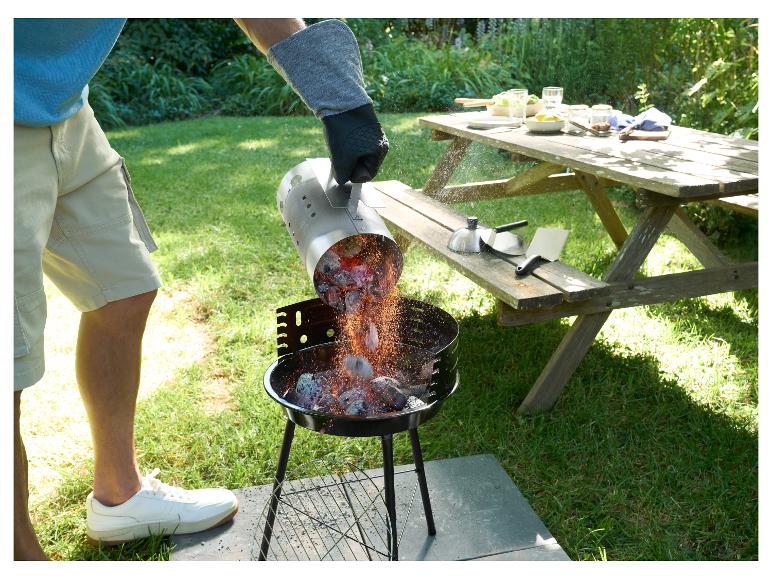 Man giet gloeiende kolen in een houtskoolbarbecue in de tuin, picknicktafel op de achtergrond.
