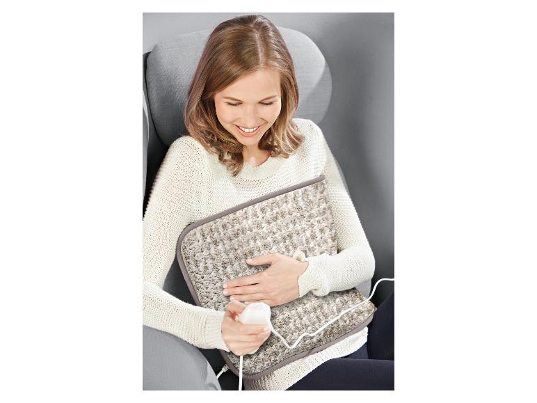 Een vrouw in een stoel glimlacht terwijl ze een beige elektrisch warmtekussen op haar schoot houdt.