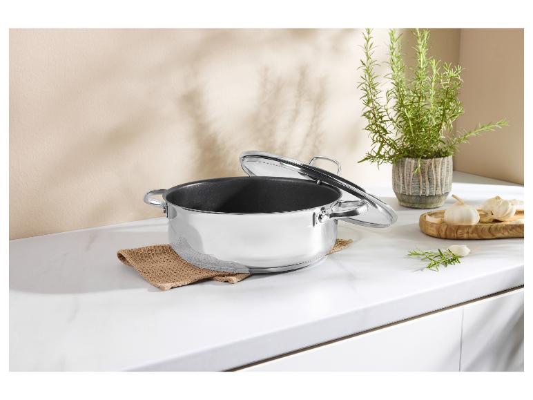 Roestvrijstalen kookpan met glazen deksel op een wit aanrecht met kruiden en knoflook.