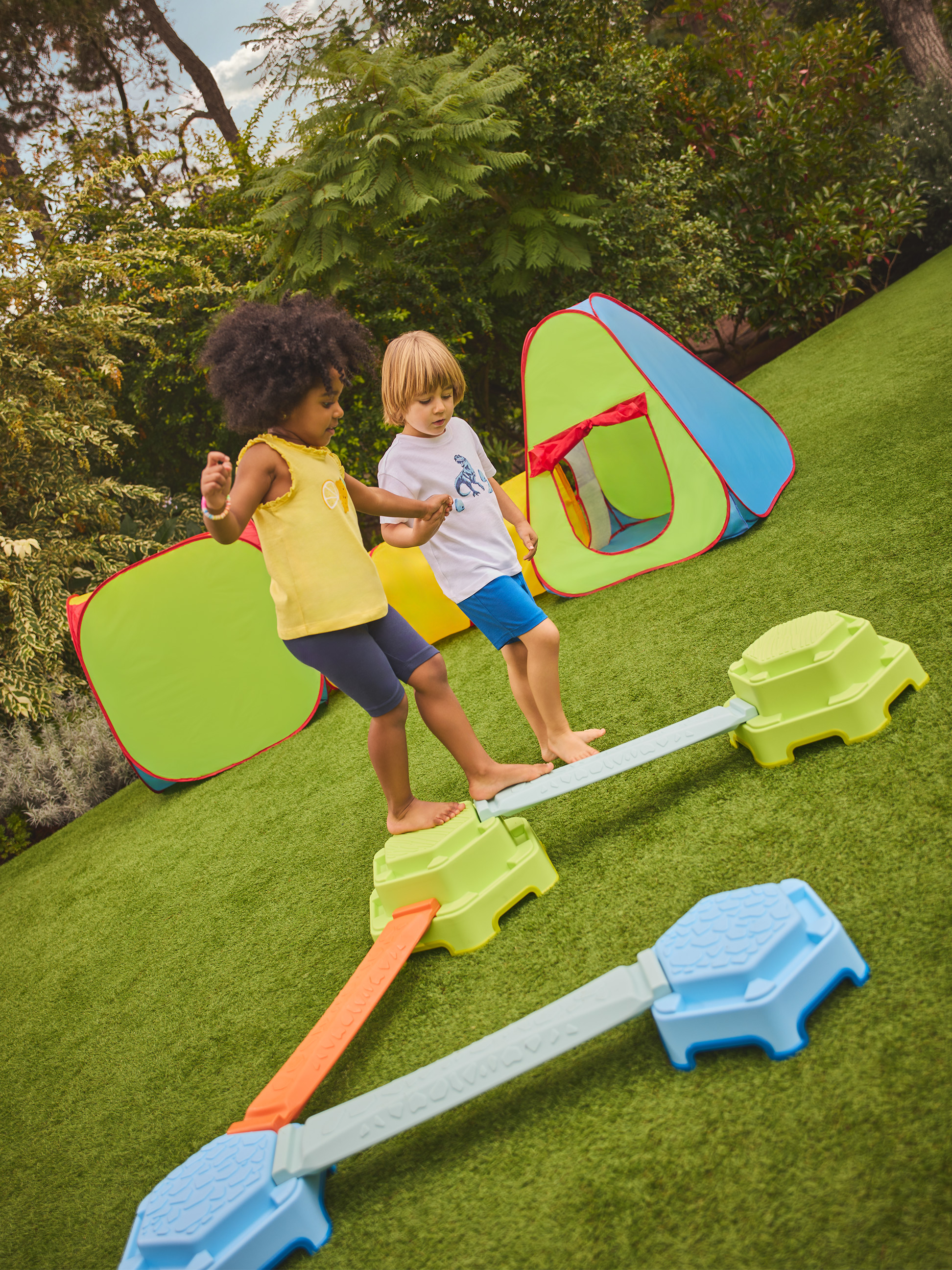 Twee kinderen spelen op een kleurrijke evenwichtsbalk met speeltenten op de achtergrond.