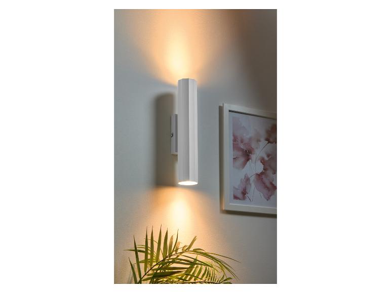 Een witte cilindrische wandlamp verlicht een kamer met een plant en een schilderij.