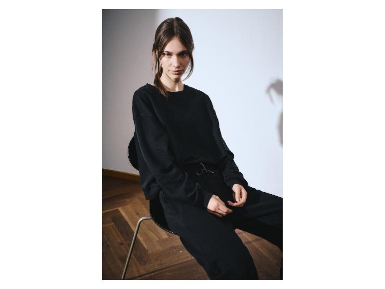 Vrouw in zwart sweatshirt en joggingbroek, zittend op een stoel.