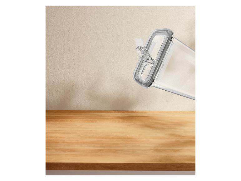 Transparante voedselbewaardoos met grijs deksel, schuin tegen een beige muur en houten oppervlak.