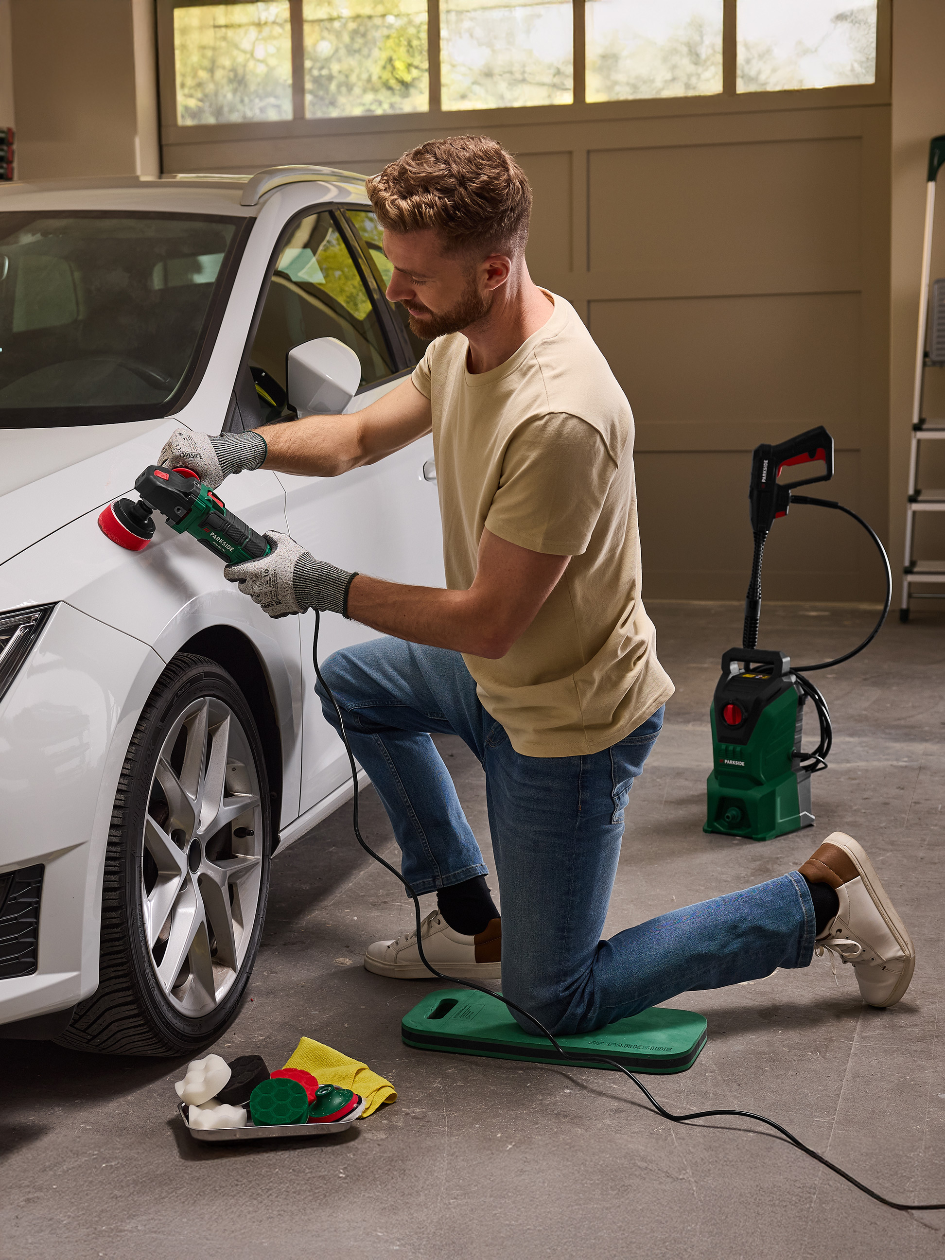 Man polijst witte auto met polijstmachine, knielend op een beschermmat.