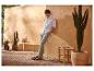 Man in wit overhemd en grijze broek, leunend tegen terracotta muur met cactussen.