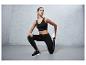 Vrouw in zwarte sportbeha, leggings en sneakers stretcht voor een grijze achtergrond.