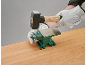 Een hand in een werkhandschoen houdt een hamer boven een klein groen aambeeld op een houten tafel.