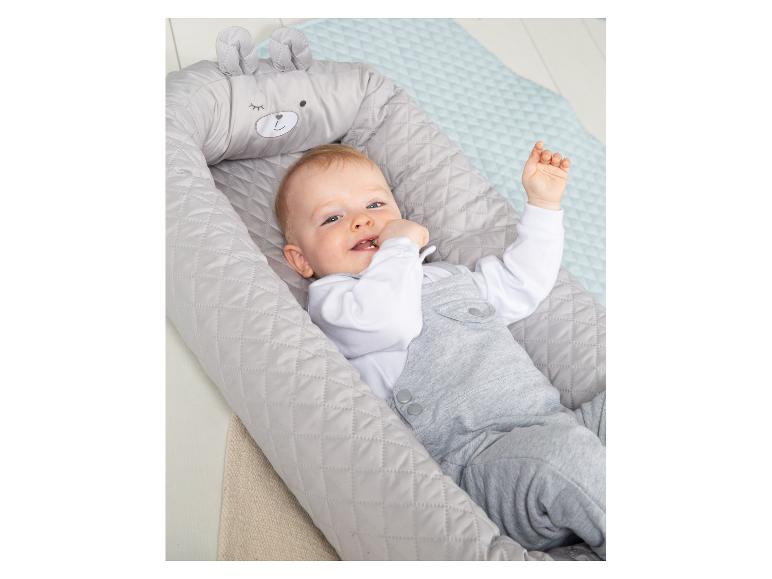 Blije baby in een grijs babynestje met berengezicht en oren, liggend op een lichtblauw kleed.