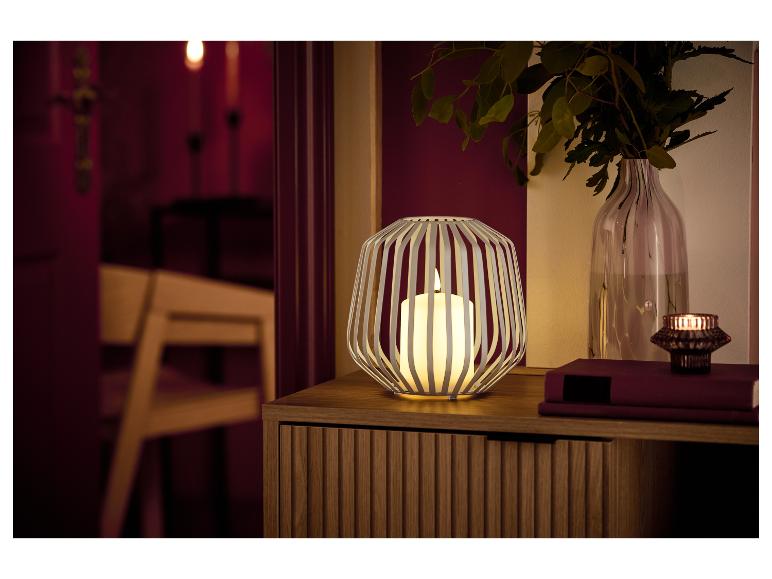 Een witte lantaarn met kaars en een vaas met groen op een houten kast.