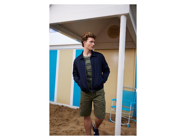Man in een marineblauwe jas, gestreept T-shirt en cargoshorts op een zandstrand.