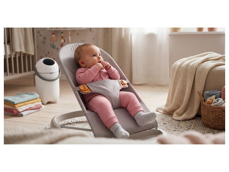 Baby in een grijze wipstoel, omringd door huishoudtextiel en een luieremmer.