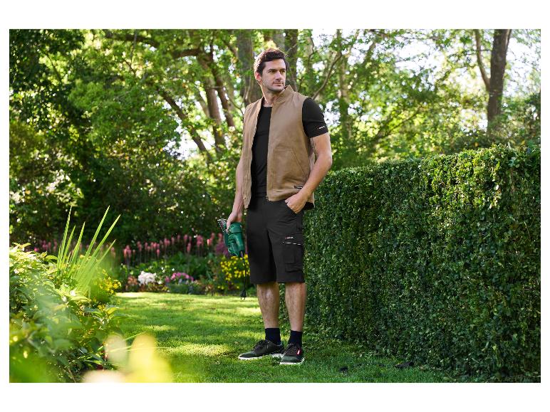 Man in bruin vest en zwarte Parkside shorts, met heggenschaar in een tuin.