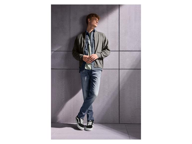 Man in grijs jack, jeans en zwarte sneakers leunt tegen een muur.