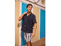 Man in een marineblauw overhemd met korte mouwen en gestreepte shorts, lachend in een strandhuisje.
