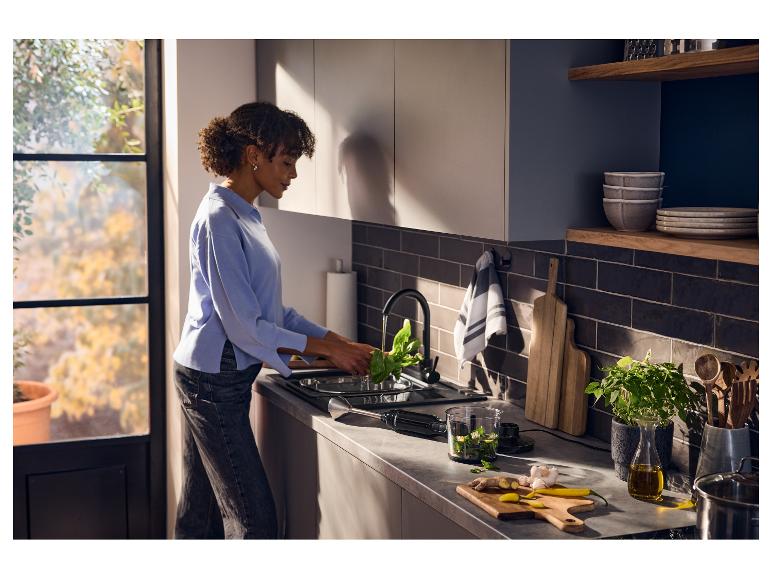 Vrouw wast spinazie in een moderne keuken met een staafmixer en ingrediënten op het aanrecht.