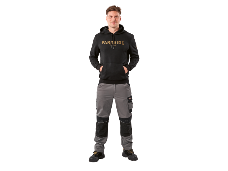 Man in zwarte hoodie en grijze werkbroek, met handen in de zakken.