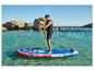 Man op blauw en rood opblaasbaar paddleboard in helder water, met rotsen op de achtergrond.