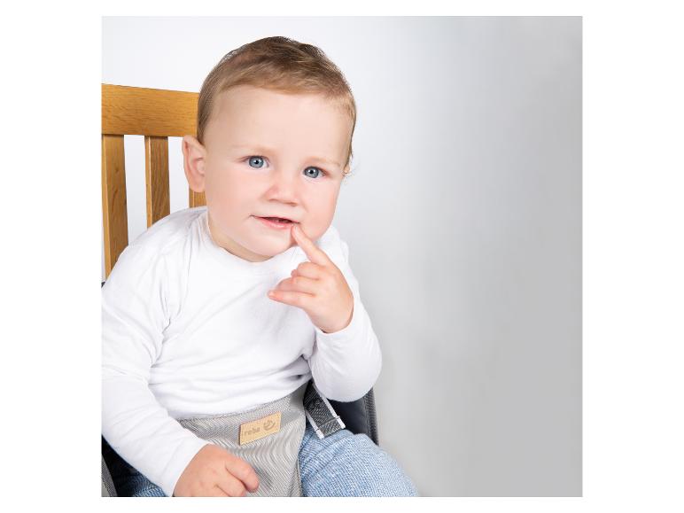 Een baby in een wit shirt en blauwe broek zit in een kinderstoel, met een vinger in de mond.