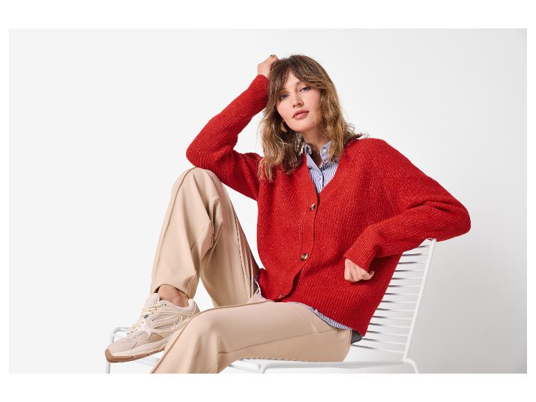 Vrouw in rood vest, beige broek en sneakers, zittend op een witte stoel.