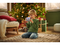 Een jongen knielt op een vloerkleed, houdt een speelgoedcamera vast, met een kerstboom en cadeaus achter hem.