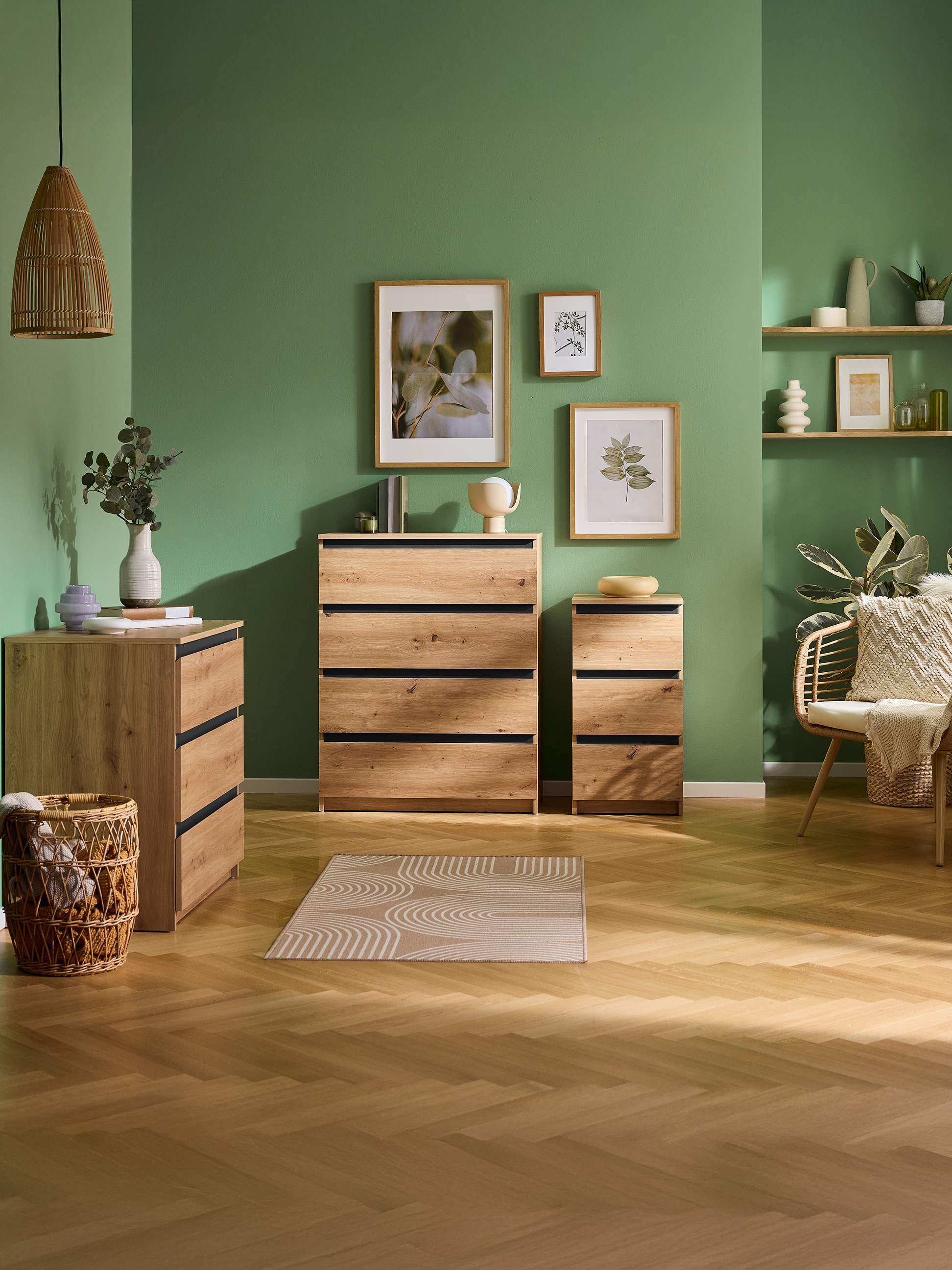 Groene kamer met houten ladekasten, planken en een rieten stoel, met botanische kunst.