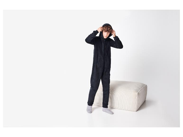 Jongen in zwart jumpsuit met capuchon en grijze sokken, staand bij een witte poef.
