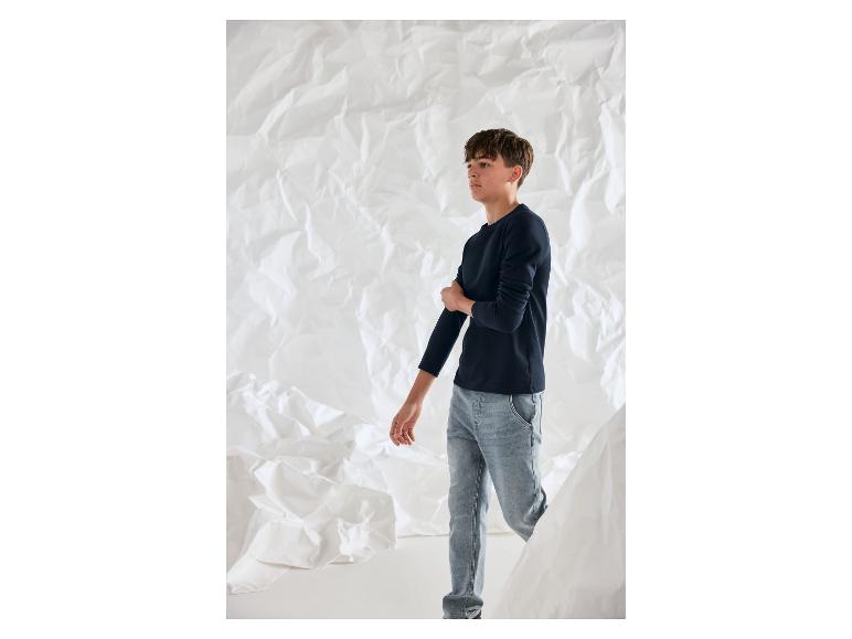 Een jongen in een donkerblauw T-shirt met lange mouwen en lichte jeans voor een gekreukelde witte achtergrond.