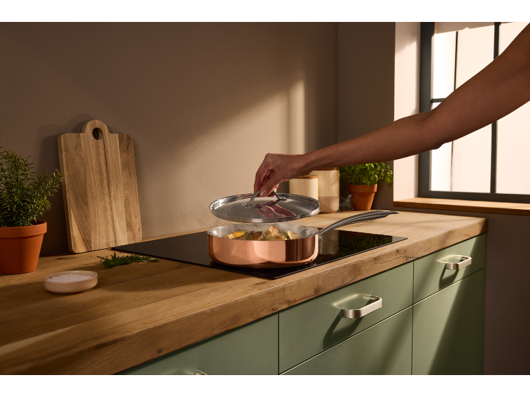 Koken in koperen pan op inductiekookplaat, met snijplank en kruiden op de achtergrond.