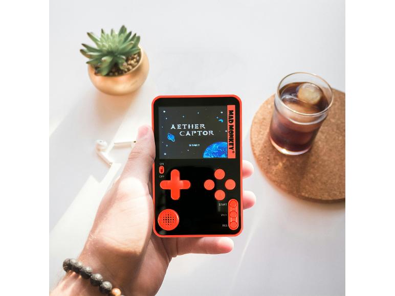 Hand houdt zwart-oranje gameconsole vast met 'AETHER CAPTOR', naast een vetplant en een drankje.