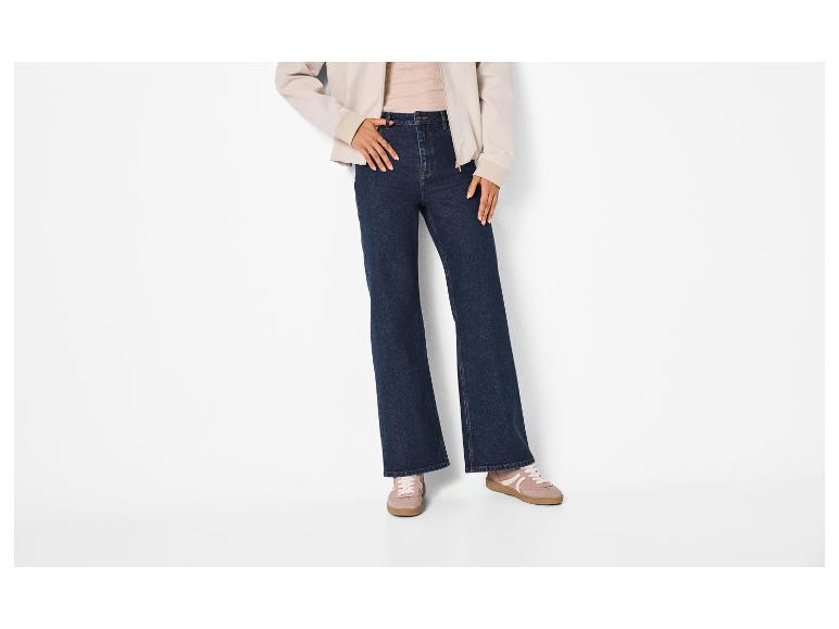 Vrouw in donkerblauwe flared jeans, beige jasje en roze sneakers