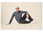 Man in grijs henley shirt, zwarte jeans en witte sneakers zit op de grond.