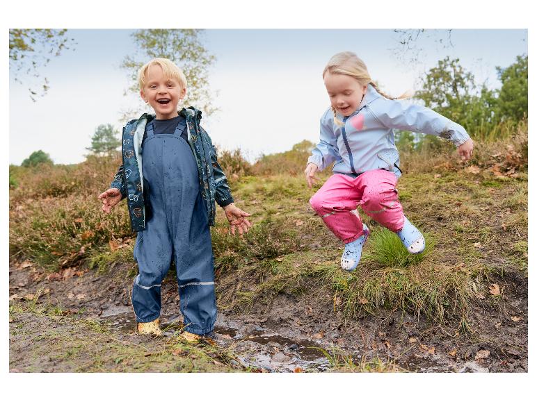 Twee blije kinderen in waterdichte jassen en tuinbroeken spelen in modderige plassen.