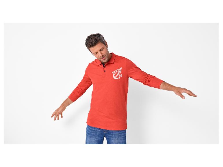 Man in een rood poloshirt met lange mouwen met ankermotief en blauwe jeans.