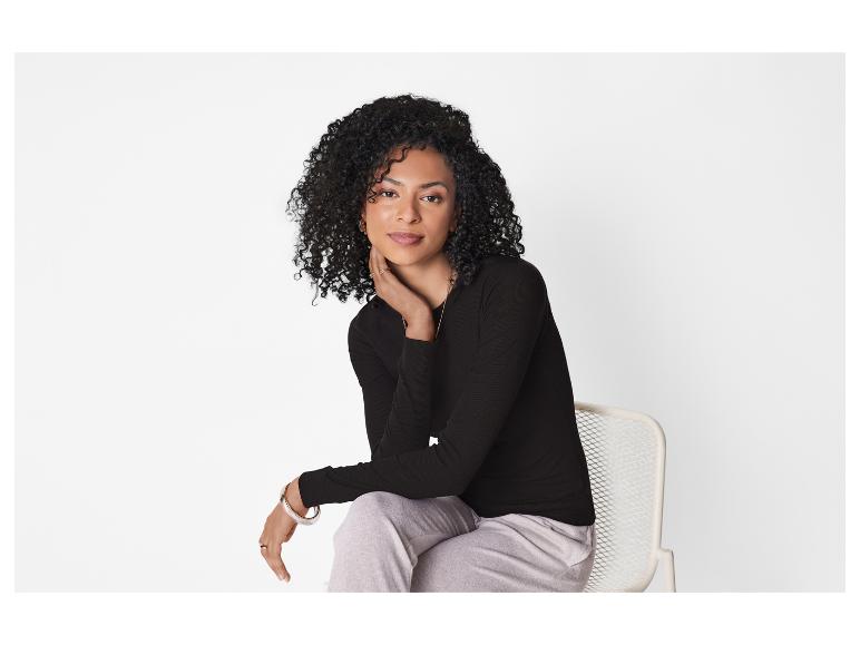 Vrouw met krullend haar, in een zwarte blouse en grijze broek, zittend op een stoel.