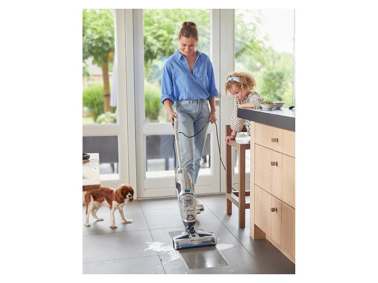 Vrouw maakt keukenvloer schoon met nat- en droogzuiger, kind en hond in de buurt.